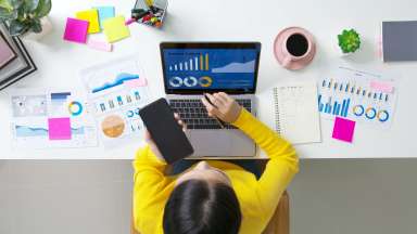 arial photo of woman looking at her computer with a screen that says &quot;business analysis.&quot; There are pages of reports on the table around the computer.