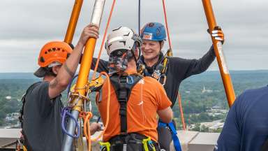 Mayor Baldwin is wearing rappelling harness and is surrounded by Over the Edge staff helping her prepare to rapel down the Wells Fargo building.