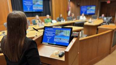 viewpoint from back of women speaking in front of a presentation on a laptop and looking at city council before her.