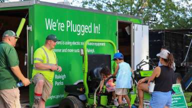 City Parks staff show members of public electric equipment with van in background that states &quot;We're Plugged in&quot;