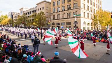 Spectators look on as marching band and flag twirlers walk down a street in downtown Raleigh