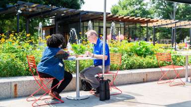 Two people are sitting at a small outdoor patio table with a flower garden behind them.