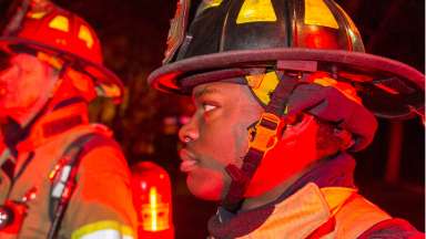 A close up of a firefighter side of face  with helmet on and chin strap. Another firefighter is in the background