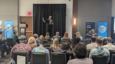 Photo of a presenter at the conference, showing them speaking on the stage and the backs of the attendees