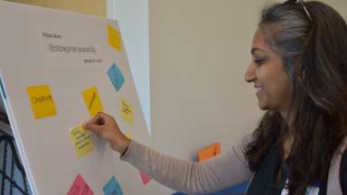 Woman smiling and placing a sticky note on a board that says &quot;What does entrepreneurship mean to you?&quot;