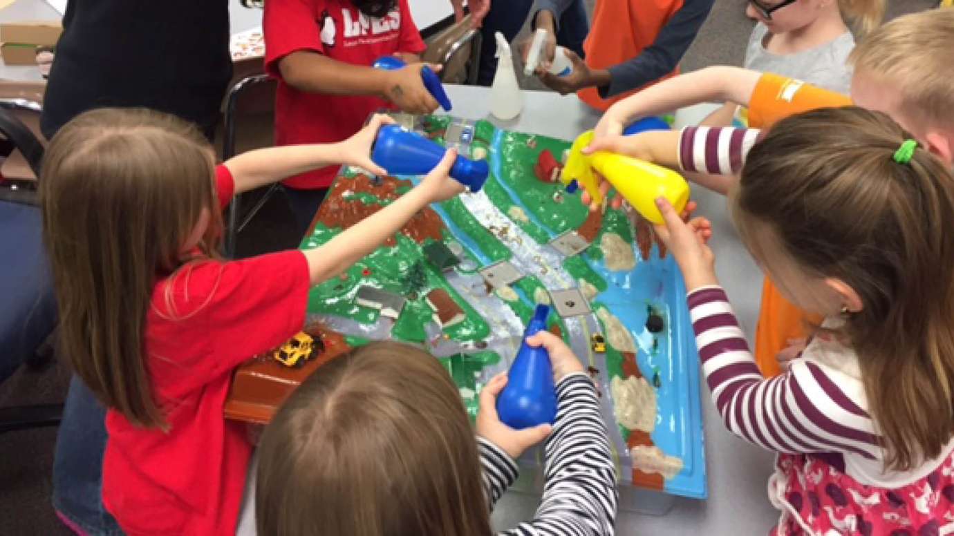 Kids spraying water on landscape roads, houses, cars, and streams to show the impact of stormwater runoff