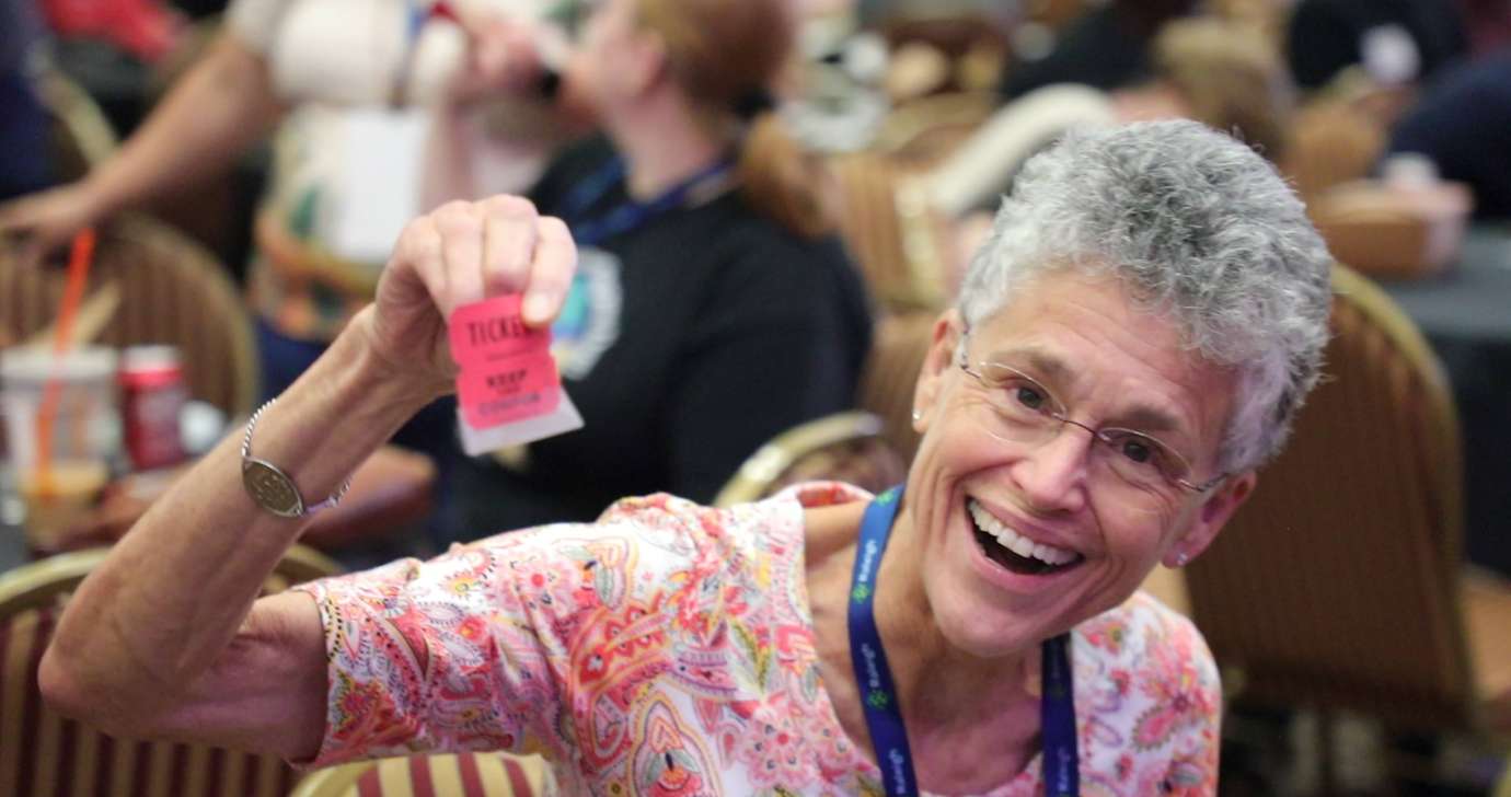 A lady with grey hair holds up her raffle ticket while smiling