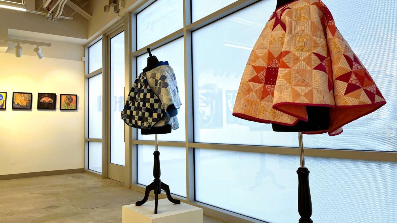 Dress forms with colorful quilts in a gallery with a row of paintings on the wall. 