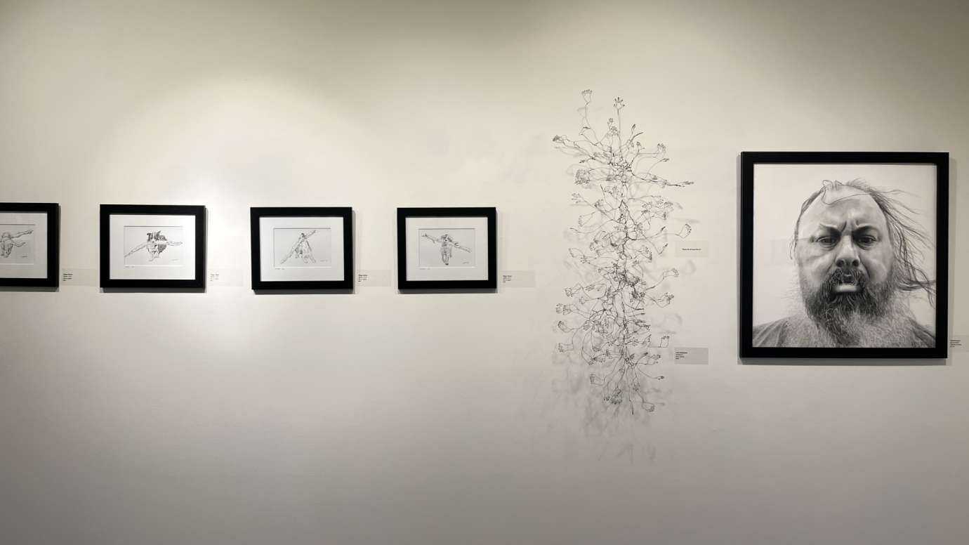 Black and white drawings in black frames on a gallery wall with a wire sculpture with hands and feet in the middle. 