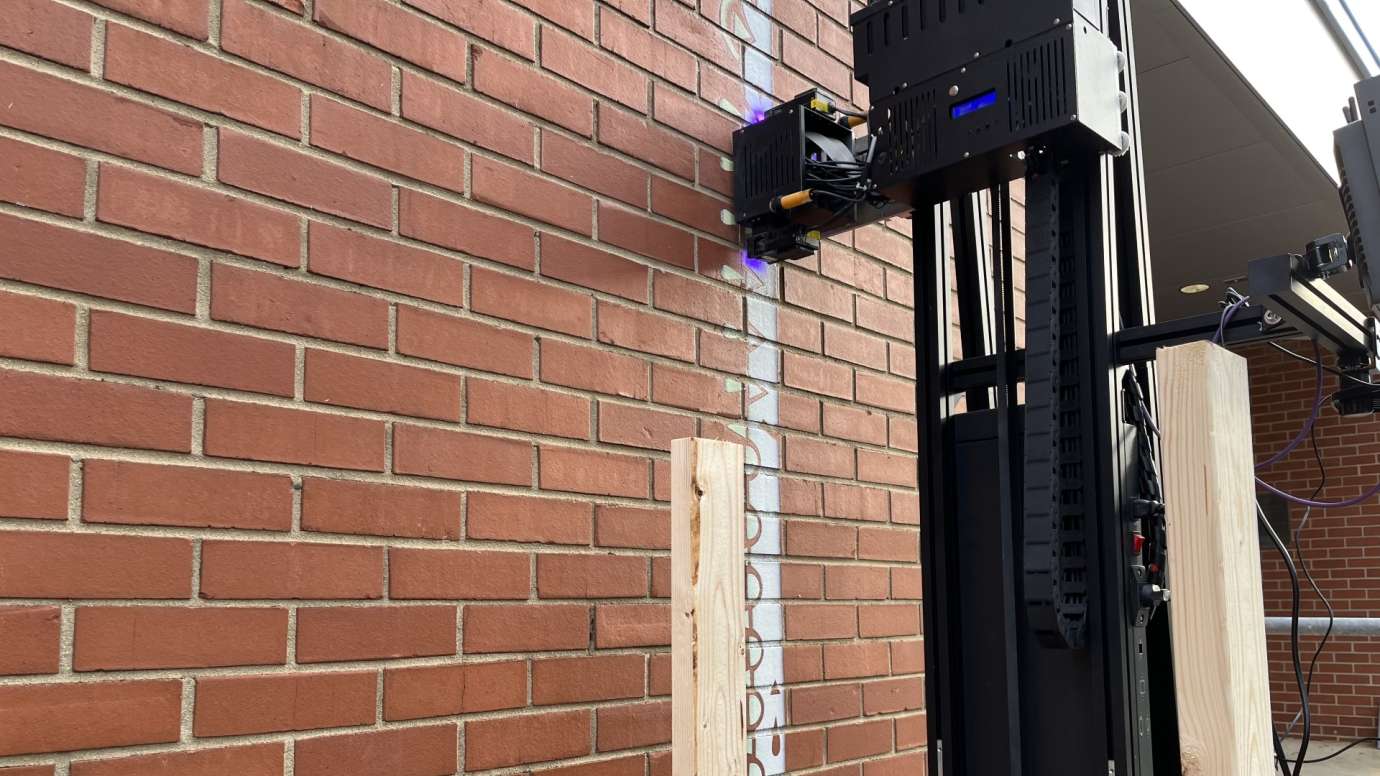 Tall black printer applying white paint to a brick wall to create a mural.