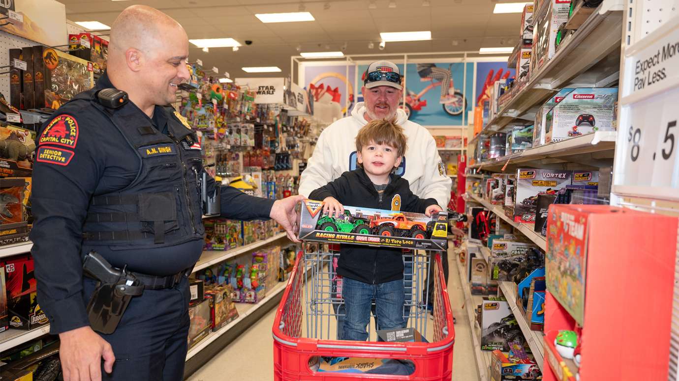 Young boy sits in a shopping cart holding a toy. Officer stands next to cart and father is pushing the cart.