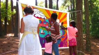 Women painting the Raleigh skyline on a banner hung between two trees