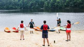 People in life vests on lake shore holding paddles during lesson