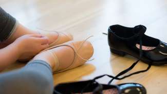 A child sitting next to tap shoes, wearing ballet shoes.