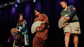 Three people dressed in costume stand on a stage holding paper fans