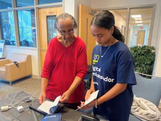 Student ambassador stands next to another person and holds the instruction booklet of a blood pressure machine which sits on the table in front of them