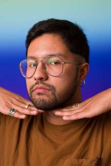 Headshot of a young latinx man with dark hair, wire aviator glasses holding his hands under his face. 