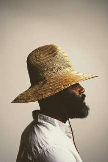 Profile of a Black man with a beard and straw hat from the shoulders up. 