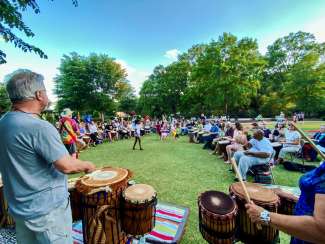 A circle of drummers sitting and playing in a grassy park area. 