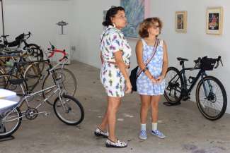 Two young women with pastel summer dresses, flowery shows and curly hair look at artwork and custom bikes in a gallery with white walls. 