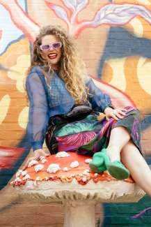 Young white woman with blond curly hair, sitting in a playful and colorful place with bright sunglasses and green shoes. 