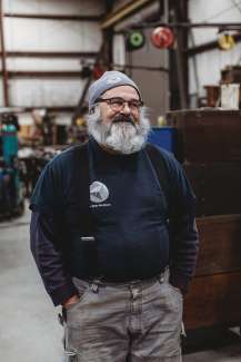 Rugged white man in a grey knit hat and white bushy heard standing in a workship with hands in his pockets.