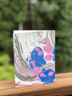 A photograph of a pink, blue, and gray marbled card against a background of trees.